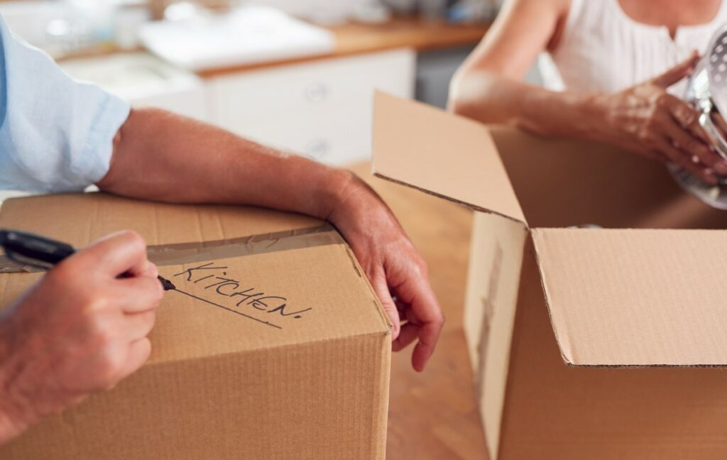 Two people label boxes as they pack, with a man writing “kitchen” on one.