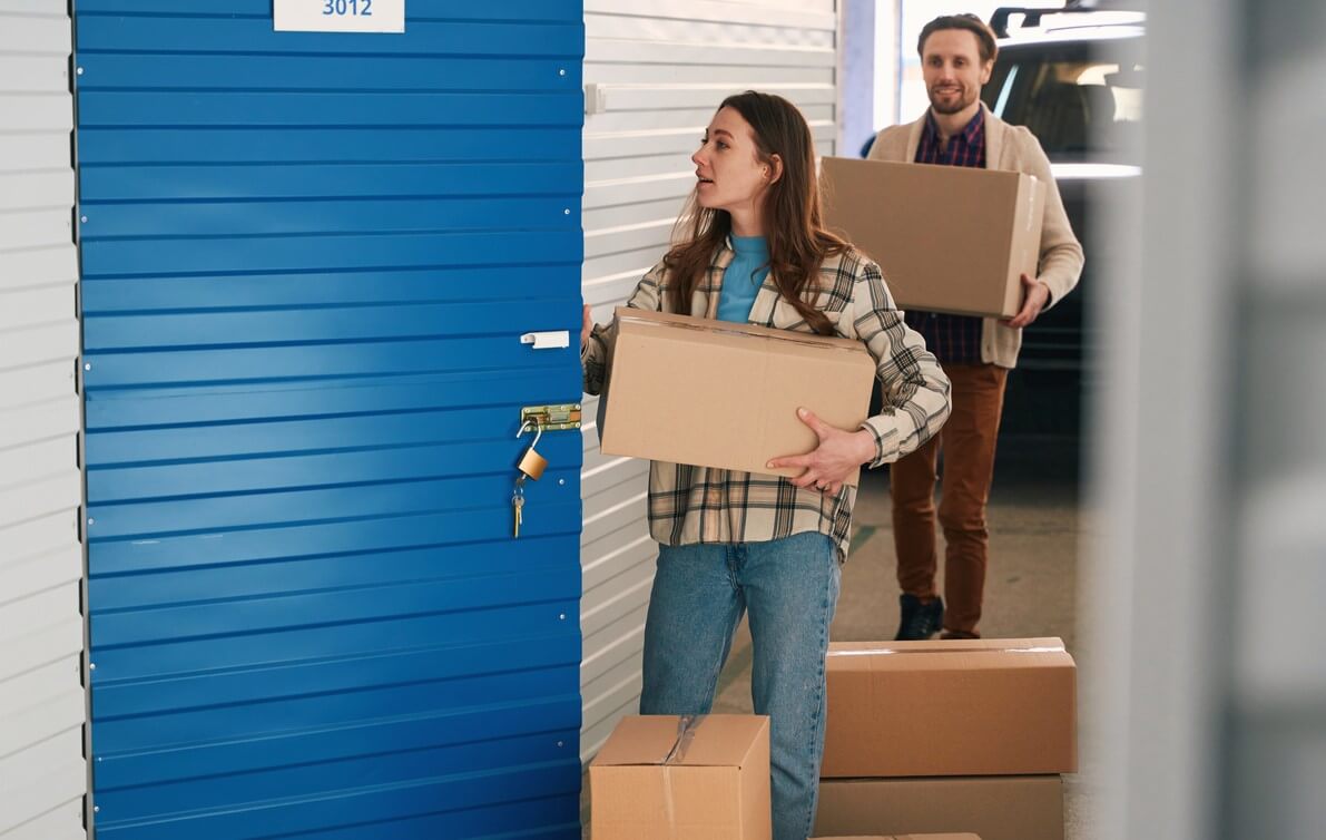 A woman and a man carry cardboard boxes into their self storage unit.