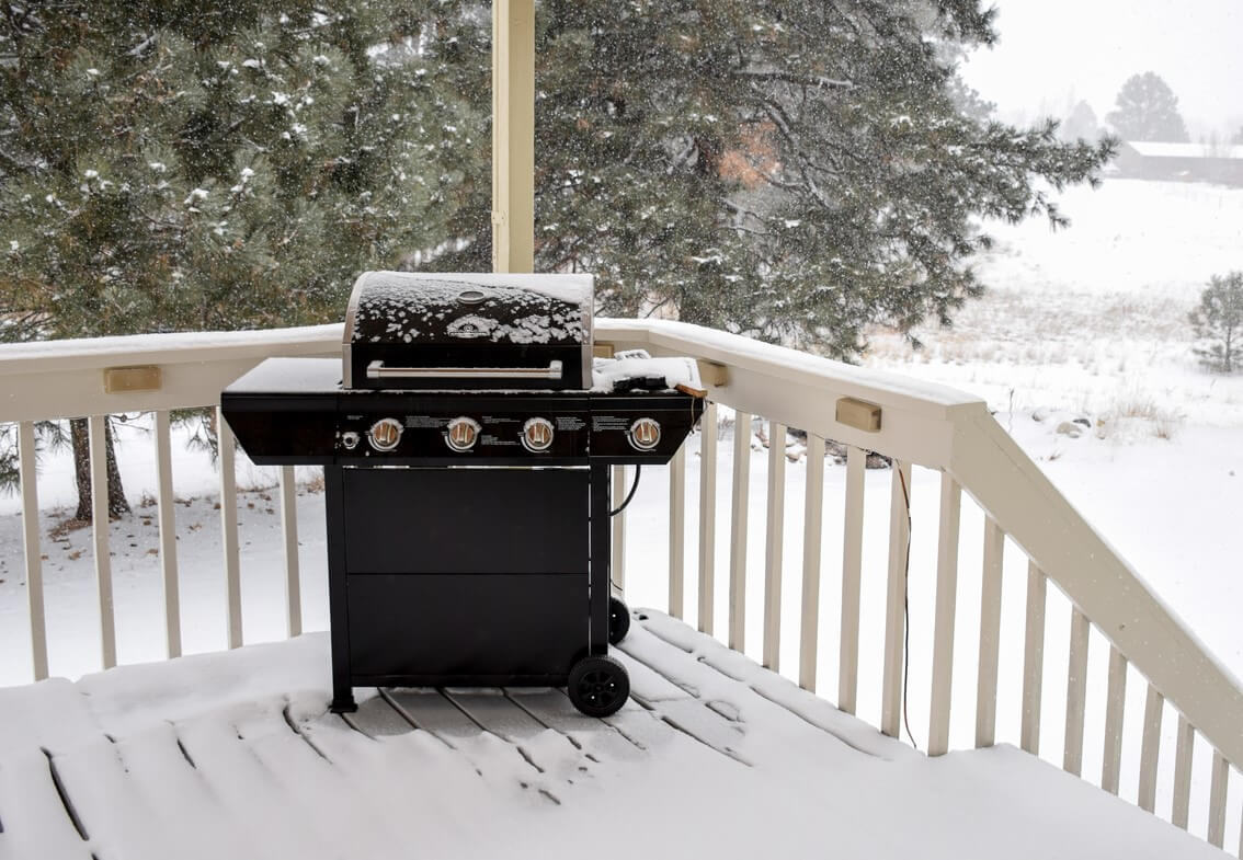 A black barbecue grill sits on a snow-covered back porch.